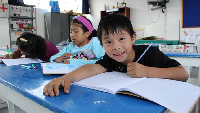 Presentación de un niño con síndrome de Down escribiendo en su cuaderno y se le ve muy sonriente .  