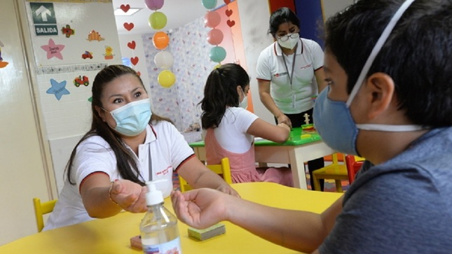Cuatro personas conversando, 2 son niños y 2 mujeres adultas. En primer plano la mujer conversa con un niño ambos tienes mascarillas. Al fondo se ve una mujer y una niña jugando.