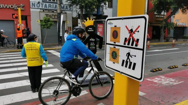 En la foto hay una persona en bicicleta y otra parada dando pase a los carros. Al costado de ambas hay un pictograma que señala en imágenes grandes los colores del semáforo  con una aspa el rojo y una fecha el color verde. Esta marcado la figura de una persona pasando .
