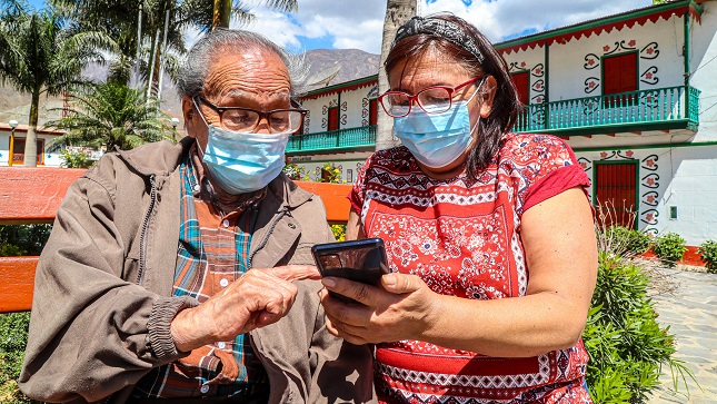 Una señora y un señor, sentados en una banca de una plaza pública de una localidad de la sierra peruana, interactúan con un smartphone.