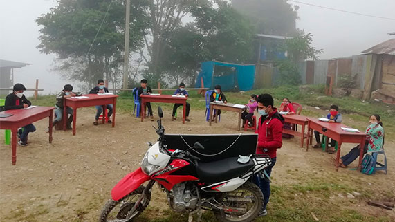 El universitario Neyber Terrones en medio de la neblina enseña a estudiantes, que están en carpetas al aire libre mientras él está junto a una pizarra que ha colocado sobre la moto