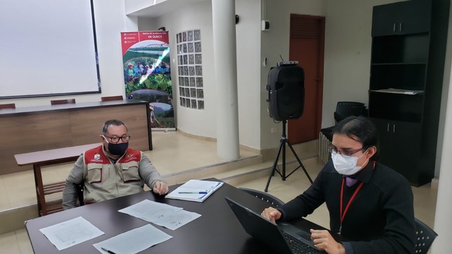 Imagen JPG de dos personas sentadas en una mesa, una de ellas revisando papeles y la otra trabajando en una laptop.