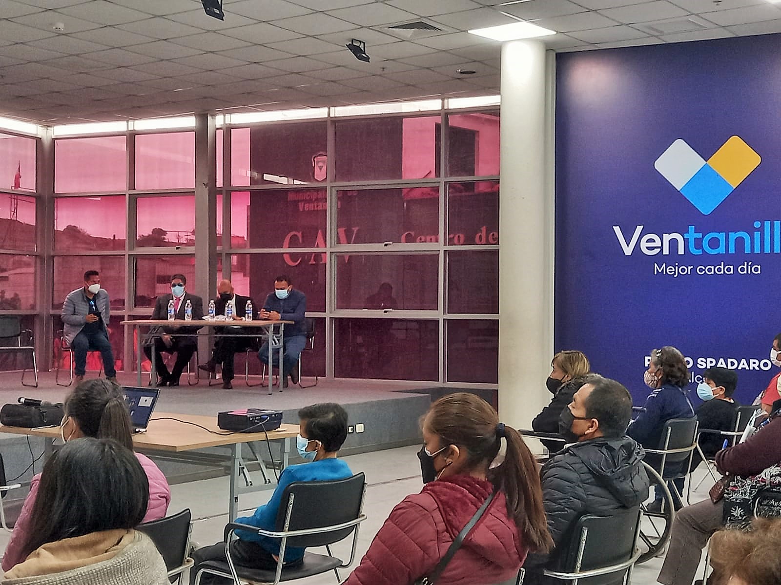 Autoridades sentadas frente a una mesa ubicada en el auditorio al fondo se ven personas escuchan la charla.