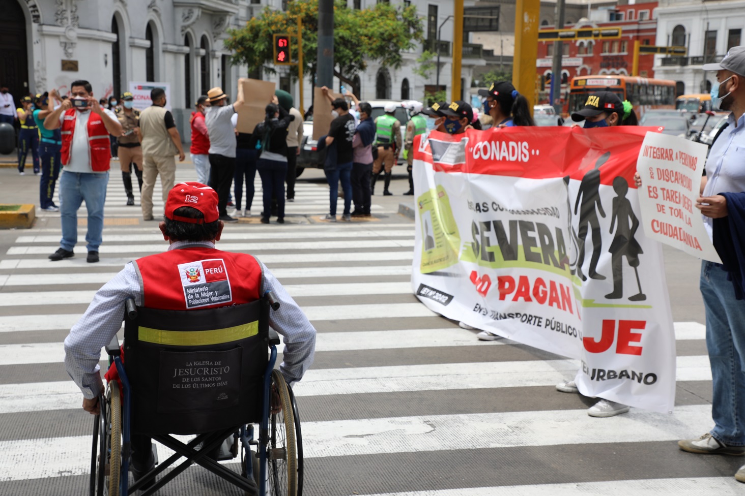 Una persona en silla de ruedas cruza una pista la fondo esta un grupo de personas con una banderola del pase libre