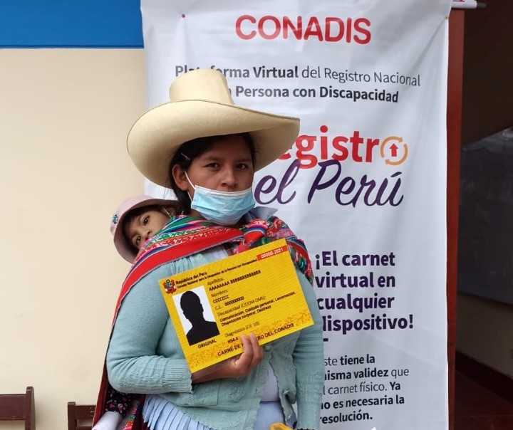 Una mujer recibe su carnet de discapacidad severa. y lo muestra esta cargada con su bebé en la espalda.