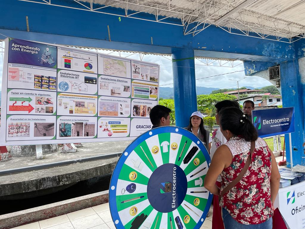 En la imagen hay 2 señoras y 3 señores hablando junto a una ruleta de electrocentro. En segundo plano hay un panel informativo del programa de aprendiendo con energía. evento realizado en de día, las personas llevan puesto ropa de verano.