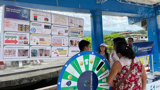 En la imagen hay 2 señoras y 3 señores hablando junto a una ruleta de electrocentro. En segundo plano hay  un panel informativo del programa de aprendiendo con energía. evento realizado en de día, las personas llevan puesto ropa de verano.
