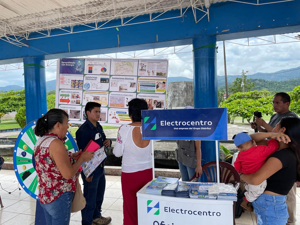 En el primer plano hay un modulo de la empresa de electrocentro y una señora con su bebe en sus brazos. En segundo plano hay 4 señores interactuando con la ruleta de electrocentro.