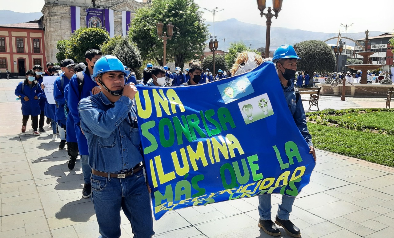 En la foto hay dos alumnos del con su uniforme jean de color azul y sus casos de color azul con un cartel con la frase: Una sonrisa ilumina más que la electricidad