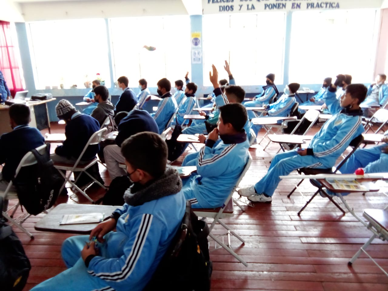 En la imagen hay un escolar levantando la mano en su salón de clases