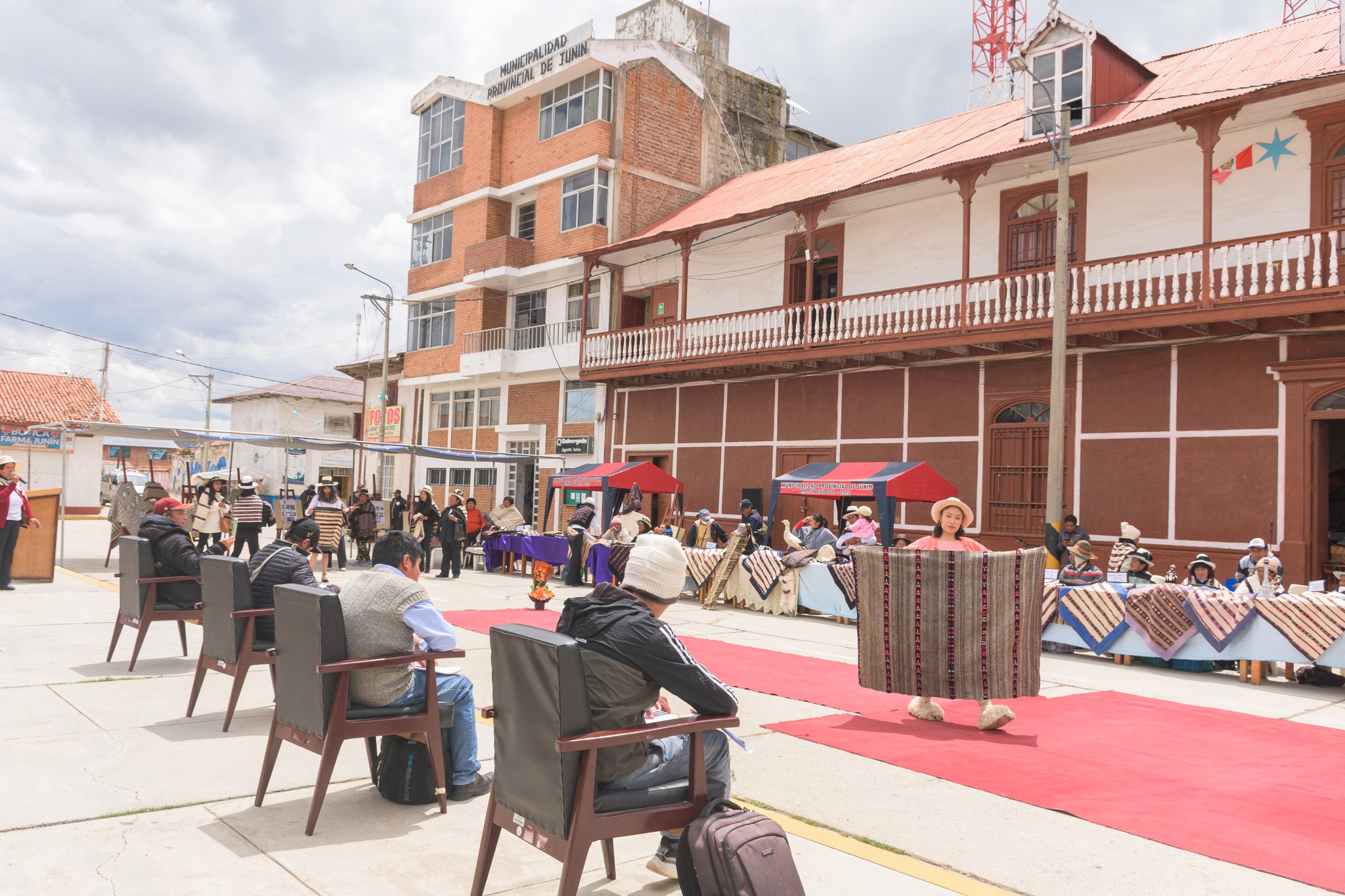 La imagen muestra el concurso de manta junina con jueces evaluando las creaciones coloridas mientras los participantes esperan con orgullo. La diversidad de colores y diseños resalta la riqueza cultural de la tradición. Una escena vibrante que captura la esencia del evento.