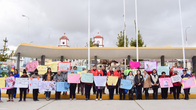 La imagen muestra mujeres con carteles en un evento de concientización, respaldadas por trabajadores municipales. Refleja la colaboración en la promoción de derechos humanos y la igualdad de género