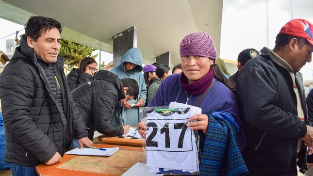 Imagen emocionante: Señora feliz sostiene su camiseta en la entrega para la Mini Maratón Junín 2023, Plaza de Armas llena de diversidad y anticipación