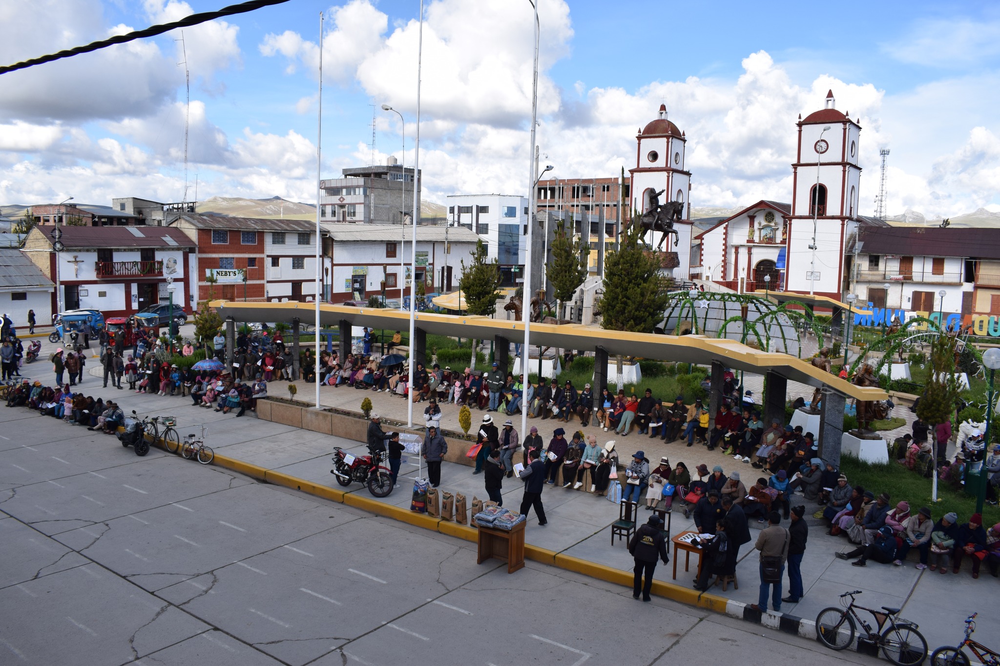 En la foto del Gran Sorteo "PREMIO AL CONTRIBUYENTE PUNTUAL 2023" en la Plaza de Armas de Junín, se ve la emoción del evento. Representantes municipales observan el sorteo, la multitud sostiene boletos con entusiasmo y los premios, como frazadas y una moto lineal, esperan ser entregados. La imagen destaca la vitalidad comunitaria y la generosidad de la Municipalidad.