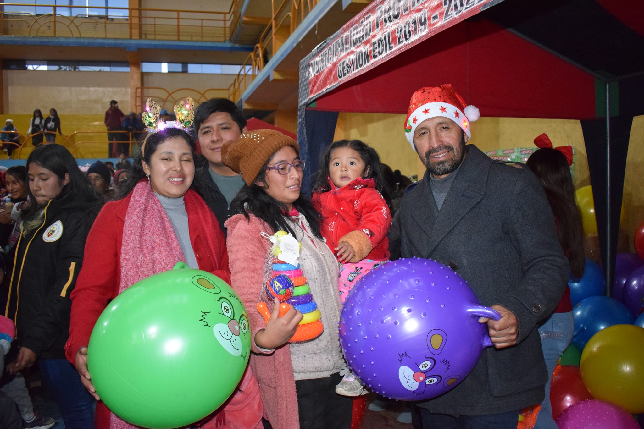 La imagen muestra al alcalde Elio Zevallos Meza sosteniendo regalos junto a una trabajadora municipal. A su lado, una madre de familia abraza a su hijo, ambos con sonrisas radiantes al recibir los regalos. La escena refleja el espíritu generoso y la conexión emocional entre la administración local y las familias durante la celebración navideña en Junín.