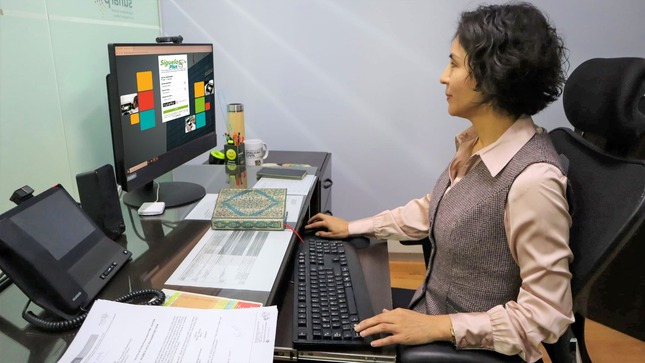 Foto de Mujer sentada frente a una computadora viendo la página web de la plataforma Síguelo Plus en el monitor