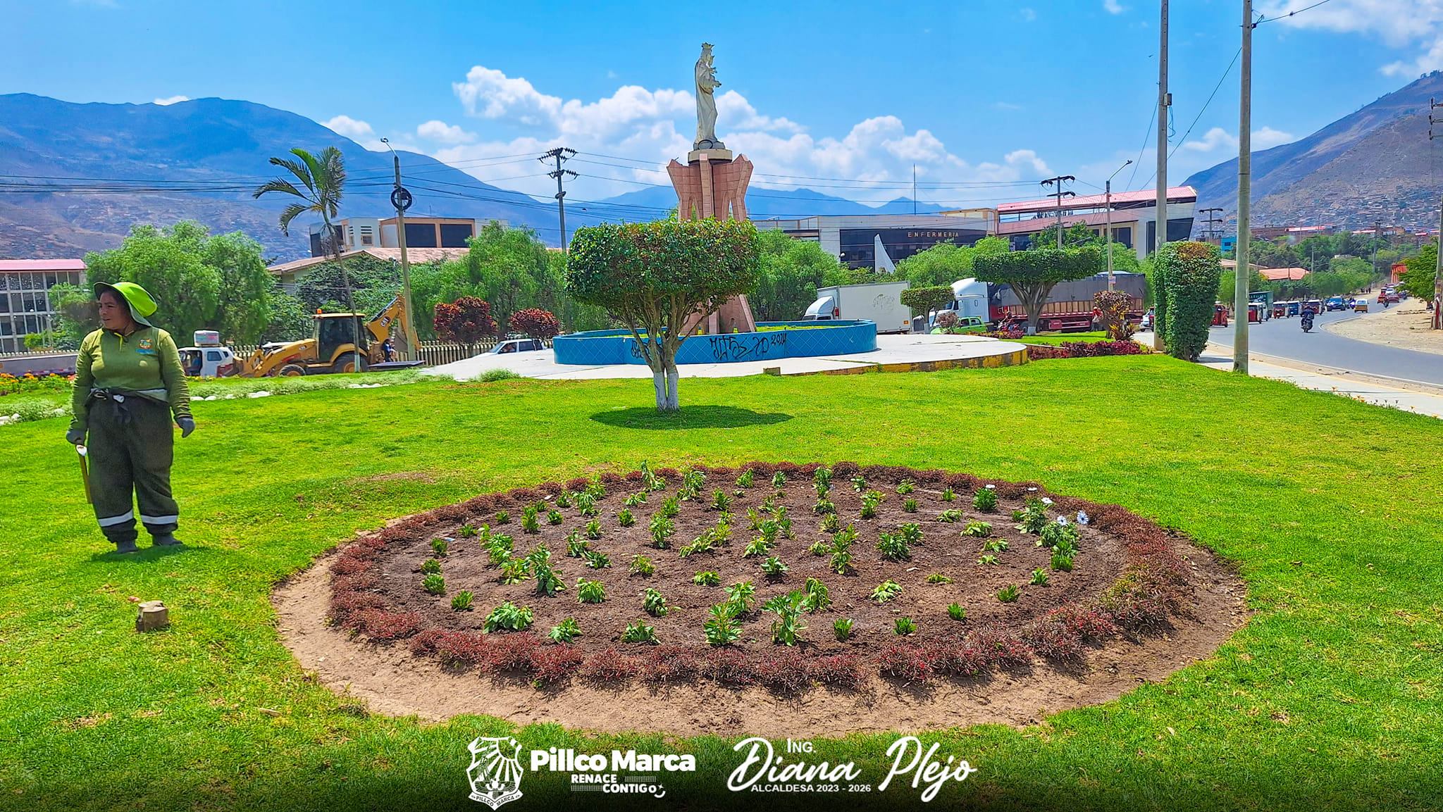 La imagen muestra un área verde con un círculo de plantas recién sembradas. Al fondo, hay una estatua elevada sobre un pedestal, rodeada por una fuente azul. También se ve una trabajadora vestida de uniforme verde, y un vehículo de construcción en el área. El paisaje incluye montañas y edificios.