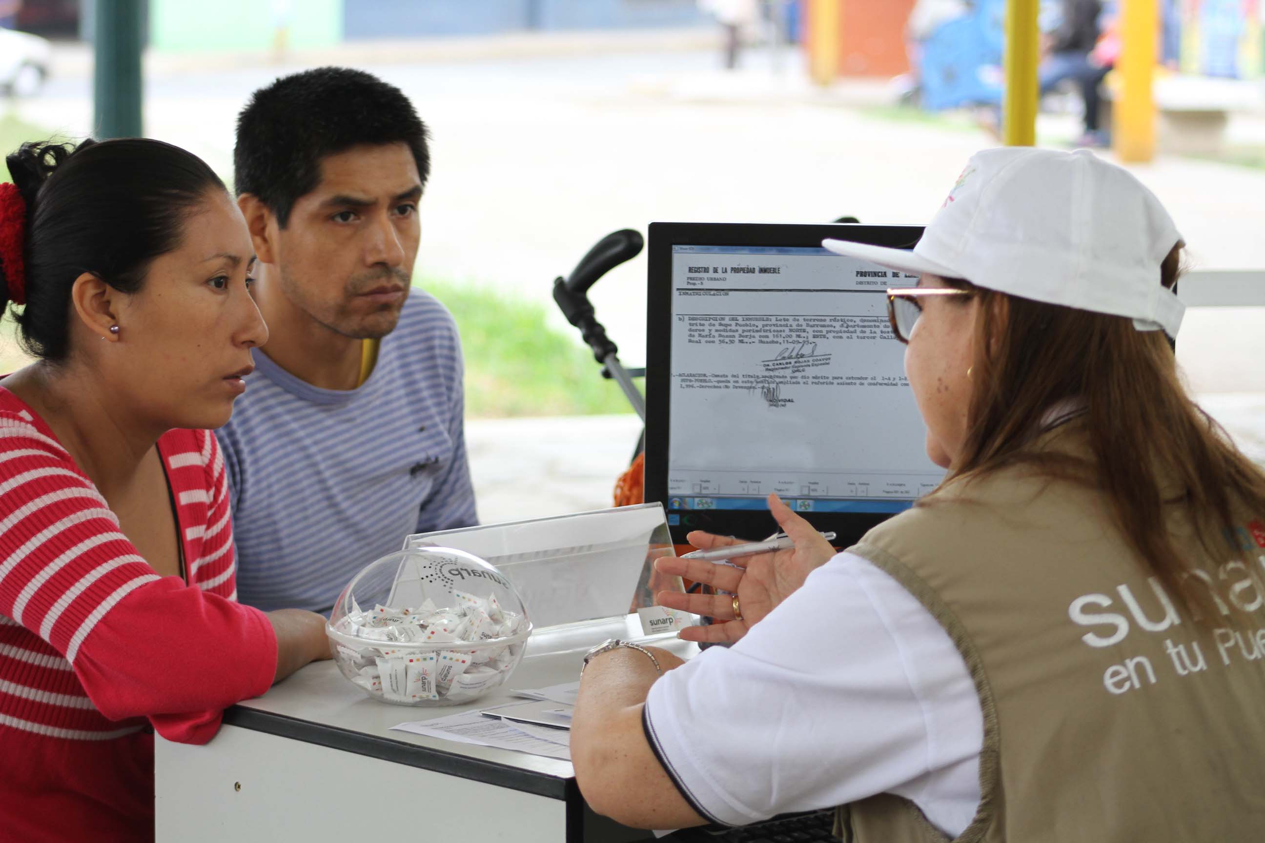 Foto de una mujer y un hombre sentados frente a módulo informativo escuchando a una colaboradora de Sunarp