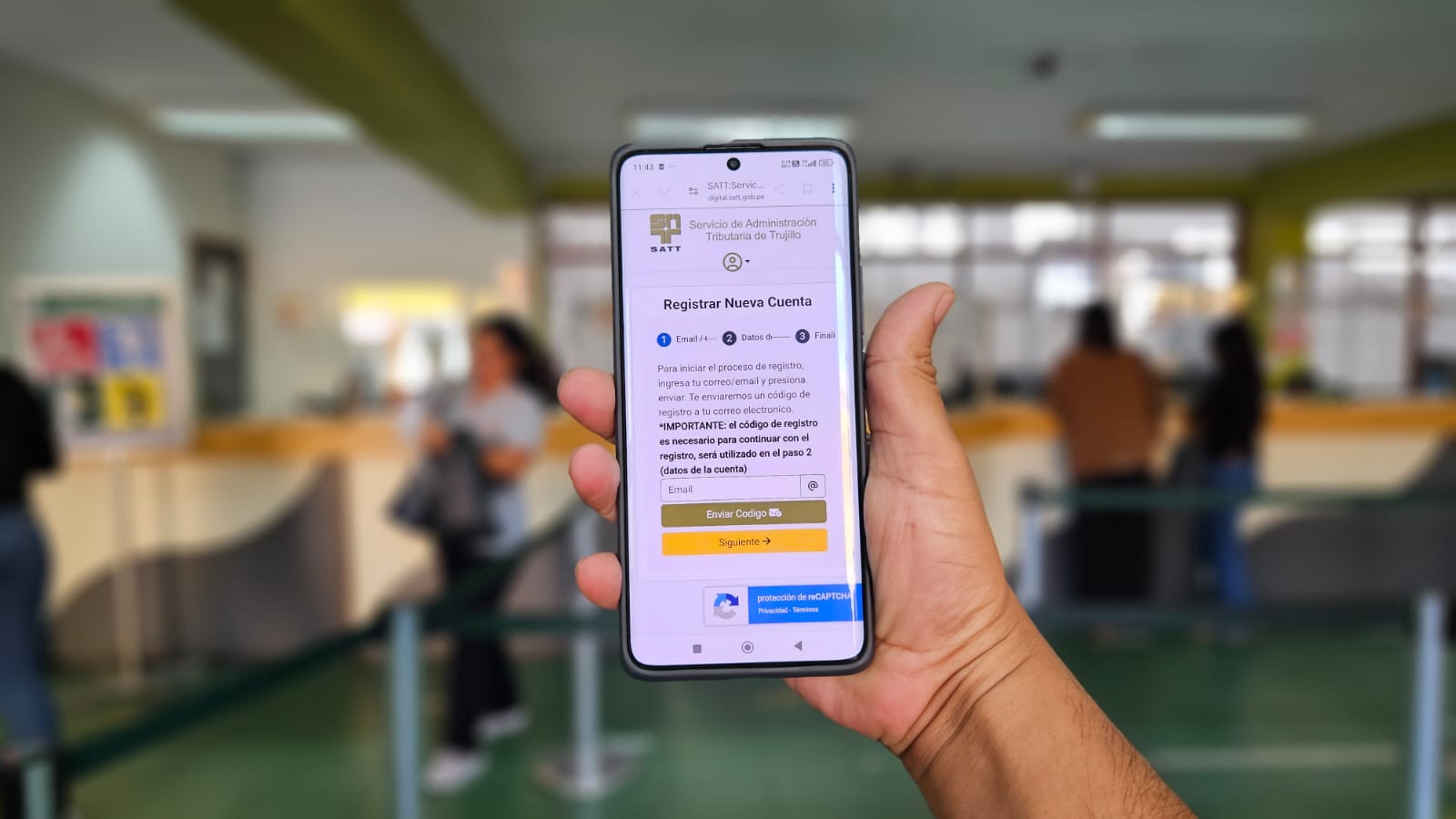 celular en mano con la página de registro a la web de pagos. De fondo las ventanillas de pago del SATT.