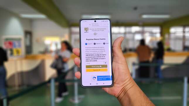celular en mano con la página de registro a la web de pagos. De fondo las ventanillas de pago del SATT.