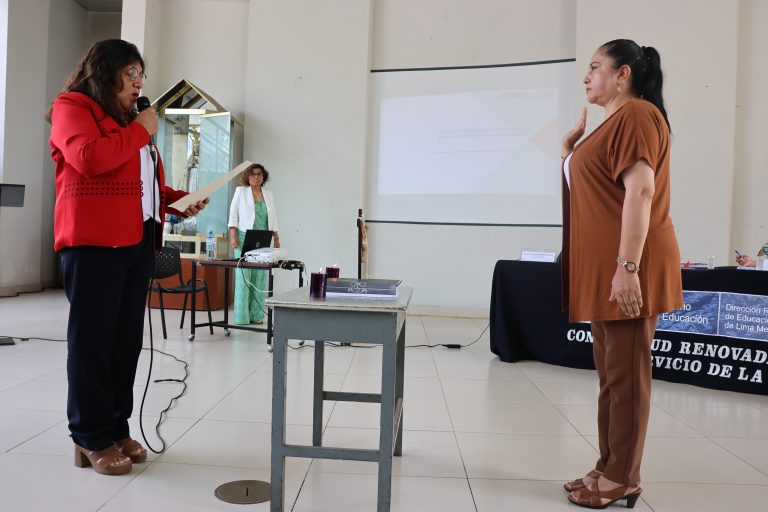 La fotografía muestra una ceremonia formal dentro de un salón institucional. Una mujer vestida de rojo sostiene un micrófono mientras lee un documento, frente a otra mujer que levanta la mano derecha, posiblemente en acto de juramentación. En el fondo se observa una pantalla de proyección y una mesa con mantel institucional.